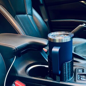 May include: A navy blue insulated tumbler with a handle sits in a black cup holder inside a car. The tumbler has a silver rim and a clear lid. The car interior features black leather seats with orange stitching.