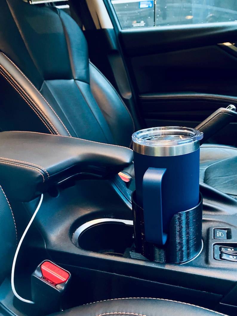 May include: A navy blue insulated tumbler with a handle sits in a black cup holder inside a car. The tumbler has a silver rim and a clear lid. The car interior features black leather seats with orange stitching.