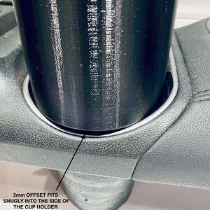 May include: A black cup holder insert, designed to fit snugly into a car's cup holder. The image shows the insert in place, with the text "2mm OFFSET FITS SNUGLY INTO THE SIDE OF THE CUP HOLDER".