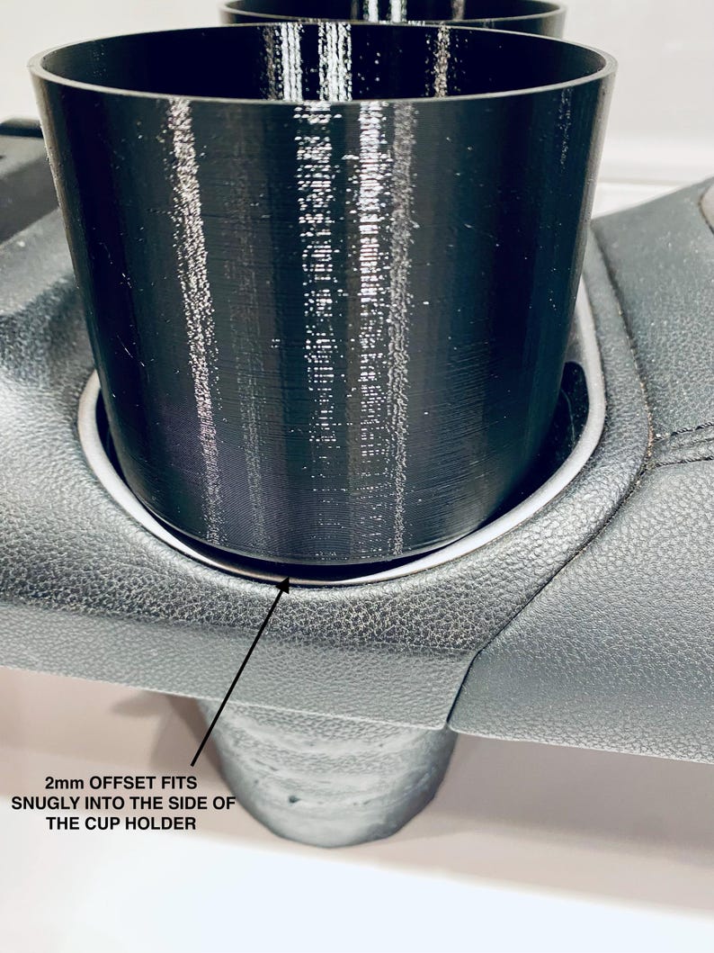 May include: A black cup holder insert, designed to fit snugly into a car's cup holder. The image shows the insert in place, with the text "2mm OFFSET FITS SNUGLY INTO THE SIDE OF THE CUP HOLDER".