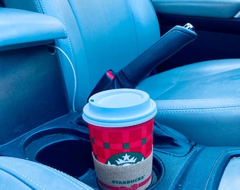 May include: A red Starbucks coffee cup with a white lid and a brown sleeve sits in a car's cup holder. The cup has a festive design with green squares and the Starbucks logo. The car interior is blue.