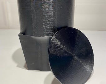 May include: A black 3D printed container with a lid. The container is cylindrical and has a square base. The lid is round and has a smooth surface.