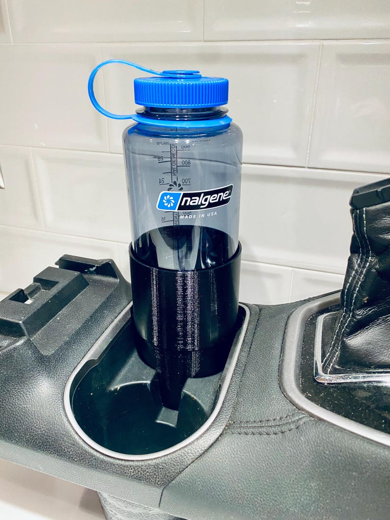 May include: A clear Nalgene water bottle with a blue lid and handle sits in a black cup holder insert. The bottle has measurement markings and the Nalgene logo. The cup holder insert is in a car's center console.