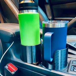 May include: A green water bottle and a blue travel mug sit in black cup holders inside a car. The water bottle has a black handle and lid. The travel mug has a blue handle. The cup holders are in the center console.