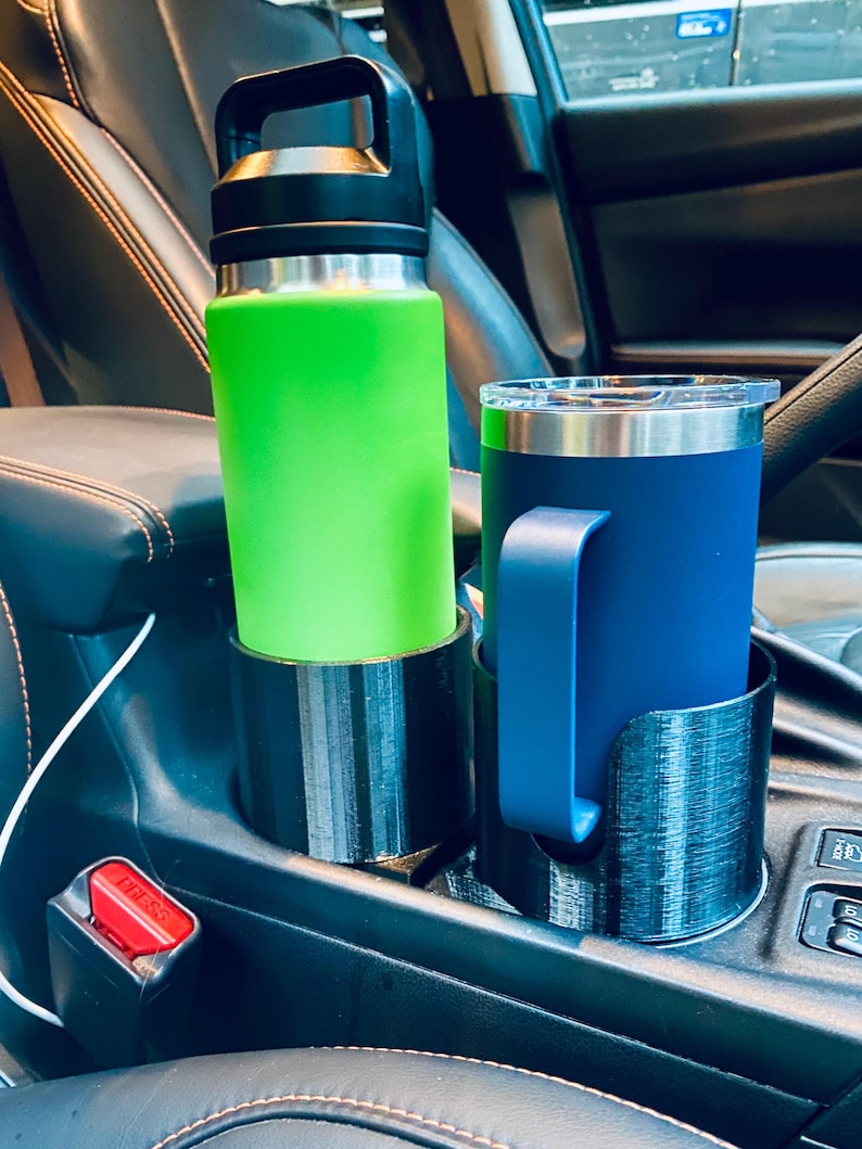 May include: A green water bottle and a blue travel mug sit in black cup holders inside a car. The water bottle has a black handle and lid. The travel mug has a blue handle. The cup holders are in the center console.