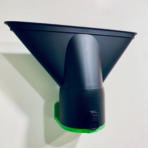 May include: A black and green vacuum attachment. The attachment has a wide, angled nozzle and a cylindrical body. The base is green, and the attachment is mounted on a white wall.