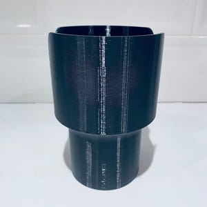 May include: A dark teal cup holder with a cylindrical base and a wider, flared top. The cup holder is made of a textured material, likely plastic, and is designed to hold a beverage container. The design is simple and functional.