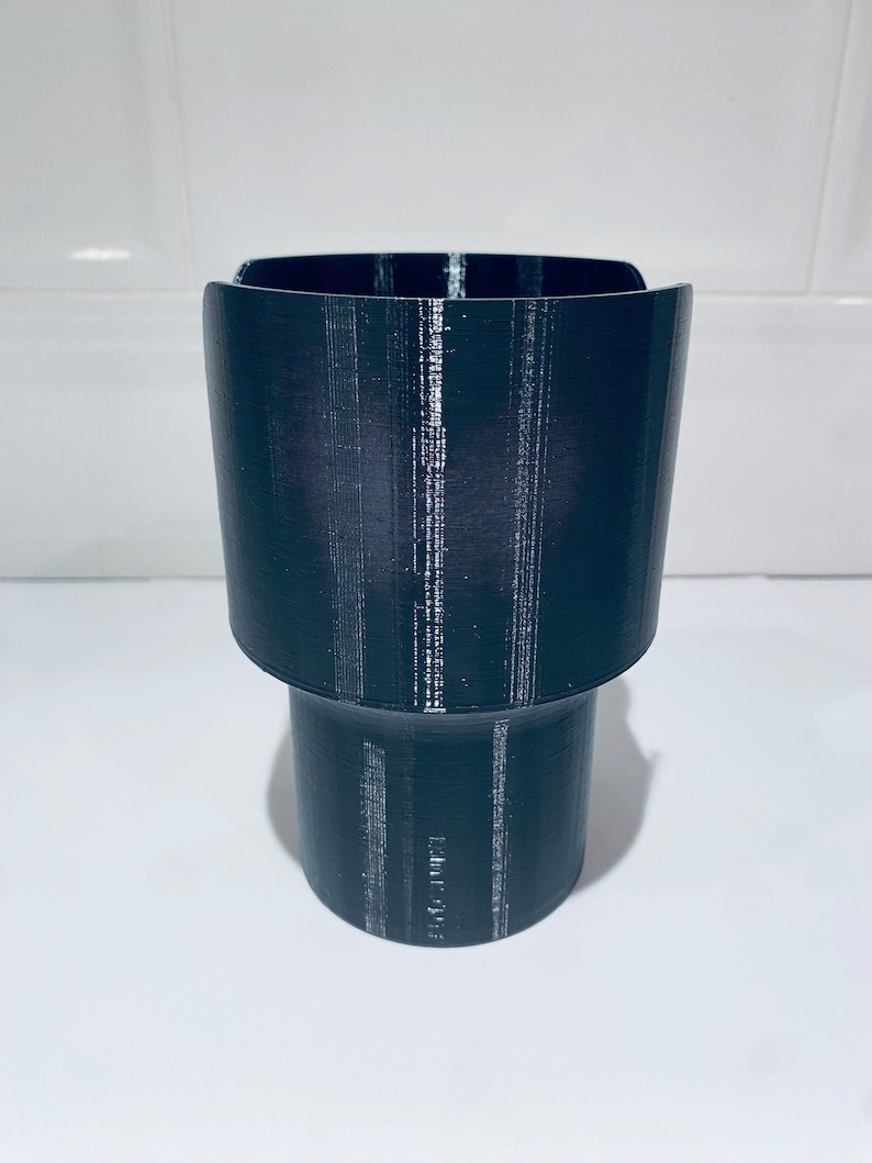 May include: A dark teal cup holder with a cylindrical base and a wider, flared top. The cup holder is made of a textured material, likely plastic, and is designed to hold a beverage container. The design is simple and functional.