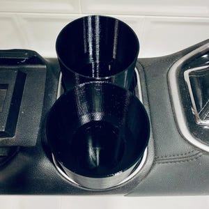 May include: Black, 3D-printed car cup holder insert. The insert features two circular cup holders of different heights, designed to fit into a car's center console. The cup holders are made of a textured plastic material and are designed to hold beverages.