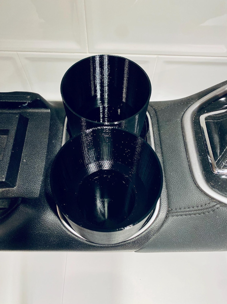 May include: Black, 3D-printed car cup holder insert. The insert features two circular cup holders of different heights, designed to fit into a car's center console. The cup holders are made of a textured plastic material and are designed to hold beverages.