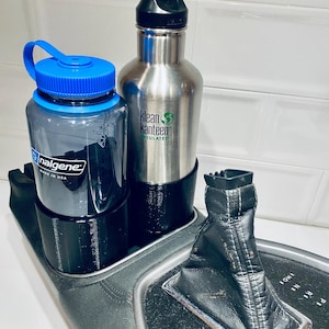 May include: Two water bottles, one clear with a blue lid and the other stainless steel, sit in a black cup holder. The stainless steel bottle has a black cap and a green logo. A black gear shift is also visible.