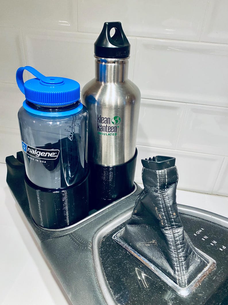 May include: Two water bottles, one clear with a blue lid and the other stainless steel, sit in a black cup holder. The stainless steel bottle has a black cap and a green logo. A black gear shift is also visible.