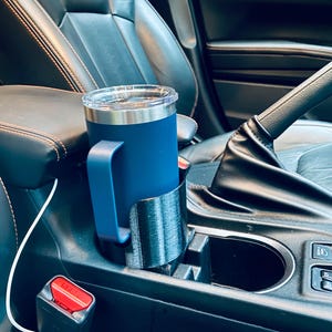 May include: A navy blue travel mug with a handle sits in a black 3D-printed car cup holder. The mug has a clear lid and is placed in the center console of a car. The car's interior is black leather.