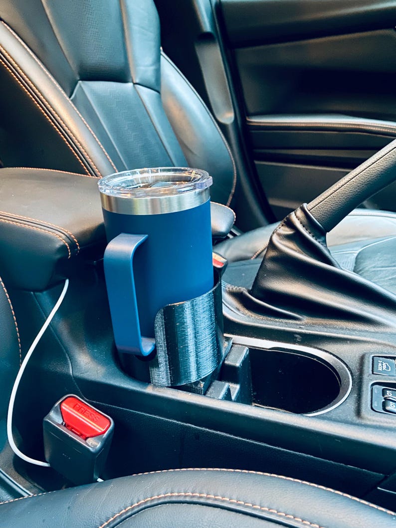 May include: A navy blue travel mug with a handle sits in a black 3D-printed car cup holder. The mug has a clear lid and is placed in the center console of a car. The car's interior is black leather.