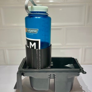 Peut inclure: Un porte-gobelet en plastique noir avec deux compartiments. Une bouteille d'eau Nalgene bleue avec un couvercle blanc est dans l'un des compartiments. La bouteille a une boucle de transport argent&eacute;e et le texte "Nalgene" et "Made in USA" imprim&eacute; dessus.
