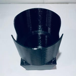 May include: A black, 3D-printed cup holder with a unique design. The holder has a wide, open top and a square base. The interior is hollow, and the exterior has a textured surface. The cup holder is designed to hold a standard-sized cup.