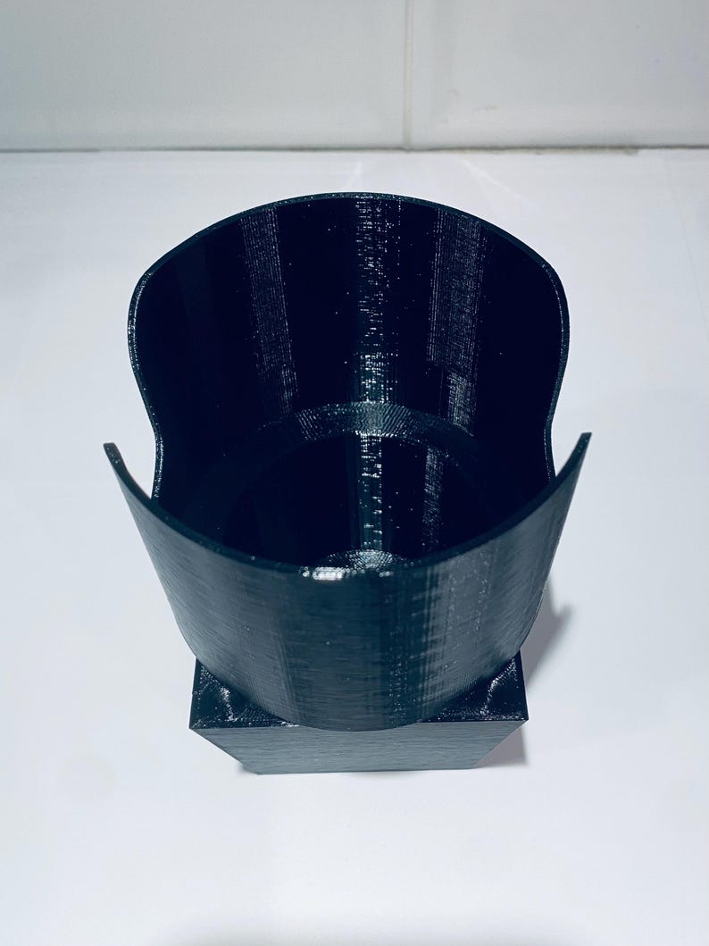 May include: A black, 3D-printed cup holder with a unique design. The holder has a wide, open top and a square base. The interior is hollow, and the exterior has a textured surface. The cup holder is designed to hold a standard-sized cup.
