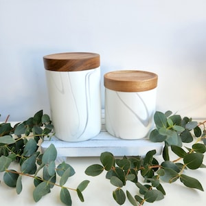 Puede incluir: Dos botes de cerámica blanca con tapas de madera. Los botes tienen un diseño de remolino gris similar al mármol. Un bote es más alto que el otro. Los botes están sobre una bandeja de madera blanca, con hojas verdes de eucalipto alrededor.