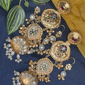 May include: Ornate gold-toned earrings with intricate detailing. Each earring features a large, circular design with embedded red, green, and clear stones, surrounded by smaller pearls. Additional pearl clusters and floral accents complete the design. The earrings are set against a dark blue background.