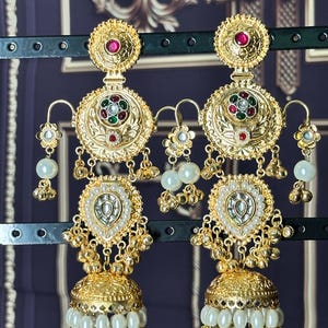 May include: A pair of gold-toned earrings with intricate detailing. Each earring features a circular top with a red gemstone, a central design with red, green, and white accents, and a teardrop-shaped pendant with white pearls. The earrings have a bell-shaped bottom with dangling pearls.