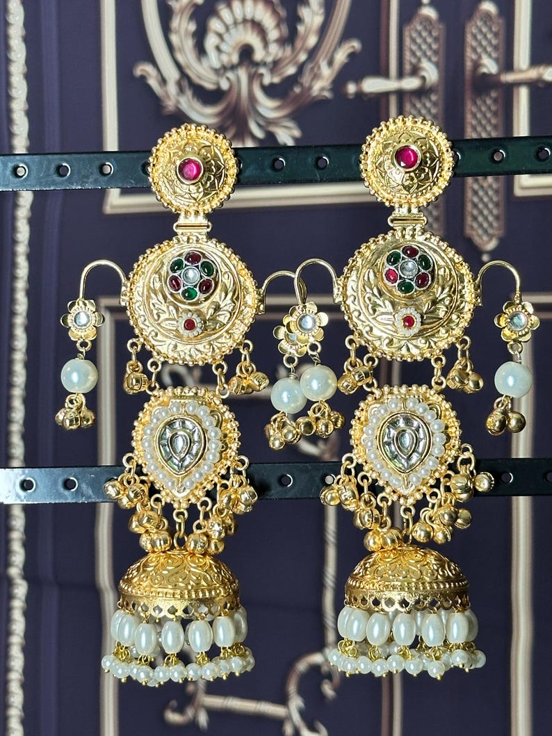 May include: A pair of gold-toned earrings with intricate detailing. Each earring features a circular top with a red gemstone, a central design with red, green, and white accents, and a teardrop-shaped pendant with white pearls. The earrings have a bell-shaped bottom with dangling pearls.