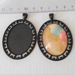 Op de afbeelding: Twee ovale hangers met sierlijke zwarte frames. Eén hanger heeft een zwart interieur, de andere heeft een kleurrijk abstract ontwerp in tinten geel, roze en blauw. De hangers zijn ongeveer 4 cm lang.