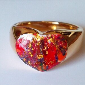 May include: A gold-colored ring with a heart-shaped centerpiece. The heart is a mix of red, white, and purple hues, accented with gold flecks. The ring has a smooth, polished finish, suitable for jewelry.