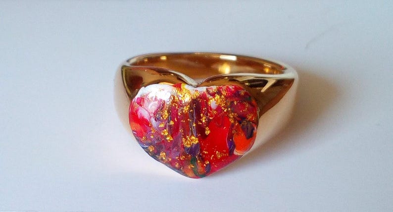 May include: A gold-colored ring with a heart-shaped centerpiece. The heart is a mix of red, white, and purple hues, accented with gold flecks. The ring has a smooth, polished finish, suitable for jewelry.