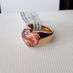 May include: A gold-colored ring with a heart-shaped centerpiece. The heart features a colorful, abstract design with red, orange, and gold accents. A white tag is attached to the ring.