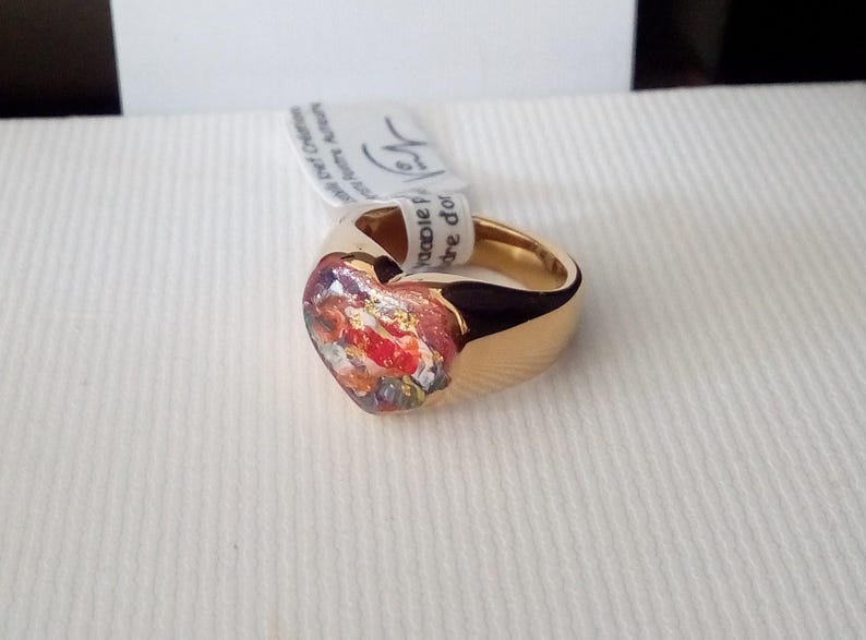 May include: A gold-colored ring with a heart-shaped centerpiece. The heart features a colorful, abstract design with red, orange, and gold accents. A white tag is attached to the ring.