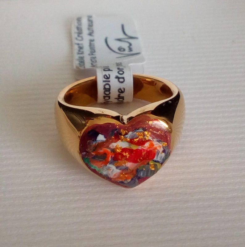 May include: A gold-colored ring with a heart-shaped face. The heart is filled with a colorful abstract painting with red, orange, blue, and gold accents. The ring has a smooth, polished finish.