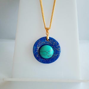 May include: A gold chain necklace with a pendant featuring a turquoise stone set in a dark blue, glitter-speckled circle. The pendant is centered on the chain, creating a striking contrast of colors and textures. The necklace is displayed on a white stand.