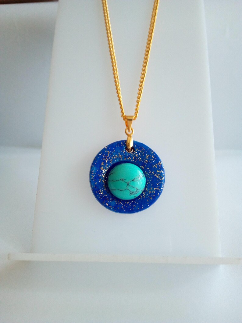 May include: A gold chain necklace with a pendant featuring a turquoise stone set in a dark blue, glitter-speckled circle. The pendant is centered on the chain, creating a striking contrast of colors and textures. The necklace is displayed on a white stand.