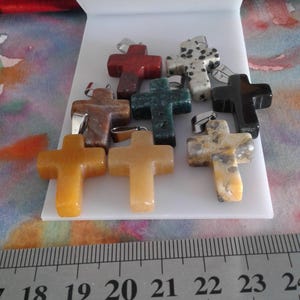 Op de afbeelding: Een verzameling edelstenen kruisbedels in verschillende kleuren, waaronder rood, groen, zwart en geel. Elk kruis heeft een zilverkleurige beugel om aan een ketting te bevestigen. De kruisen worden op een wit oppervlak getoond.