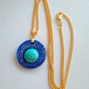 May include: A gold-colored chain necklace with a pendant. The pendant is a dark blue circle with gold glitter and a turquoise stone in the center. The necklace is displayed on a white background.
