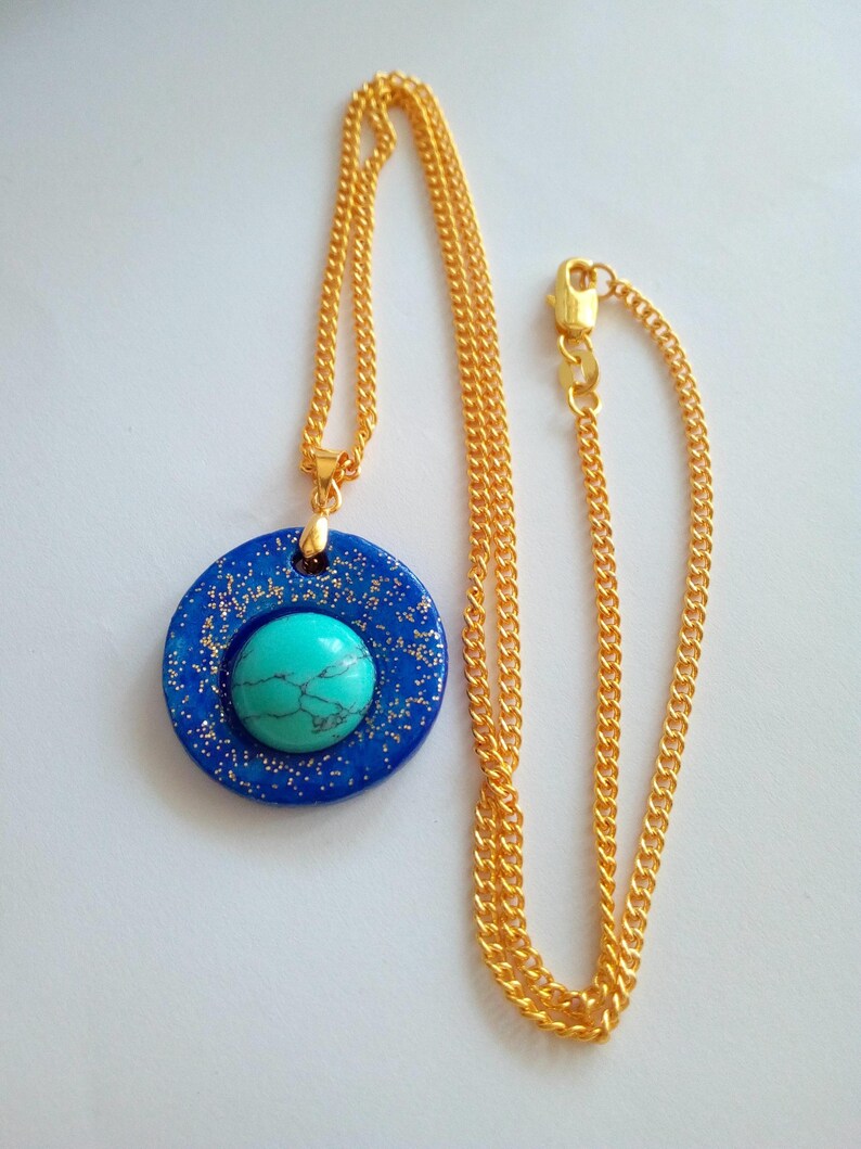 May include: A gold-colored chain necklace with a pendant. The pendant is a dark blue circle with gold glitter and a turquoise stone in the center. The necklace is displayed on a white background.