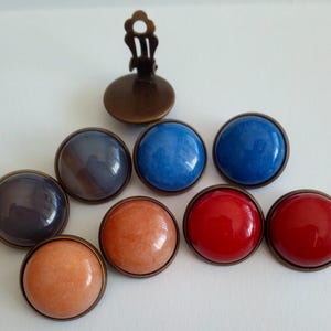 Peut inclure: Collection de boucles d'oreilles à clipser de différentes couleurs, dont le bleu, le rouge et l'orange. Chaque boucle d'oreille présente une pierre ronde et brillante sertie dans un cadre en métal de couleur bronze. Le mécanisme à clip est également visible.