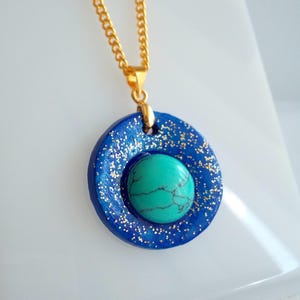 May include: A gold chain necklace with a blue pendant. The pendant has a circular shape with a turquoise stone in the center. The blue circle is decorated with gold glitter.