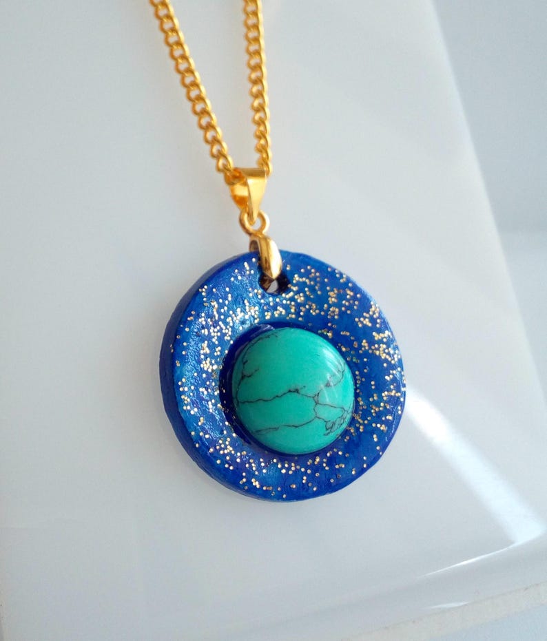 May include: A gold chain necklace with a blue pendant. The pendant has a circular shape with a turquoise stone in the center. The blue circle is decorated with gold glitter.