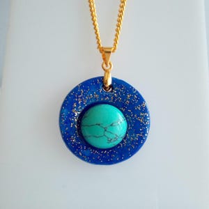 May include: A gold chain necklace with a pendant. The pendant features a turquoise stone set in a dark blue, circular frame with gold glitter accents. The necklace is a jewelry item.