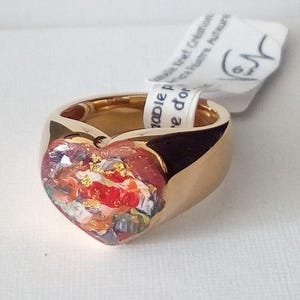May include: A gold-colored ring with a heart-shaped centerpiece. The heart is filled with a colorful, abstract design featuring red, white, and gold accents. A white tag is attached to the ring.