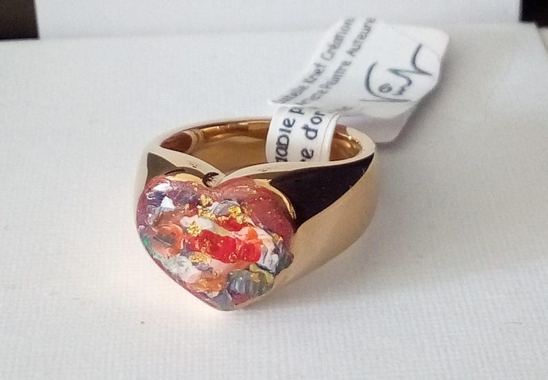 May include: A gold-colored ring with a heart-shaped centerpiece. The heart is filled with a colorful, abstract design featuring red, white, and gold accents. A white tag is attached to the ring.