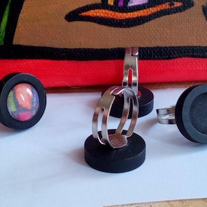 Peut inclure: Trois bagues noires avec des bandes métalliques réglables. Deux bagues ont un bézel rond noir avec un insert rond en verre transparent. Une bague a un bézel rond noir avec un insert en tissu coloré.