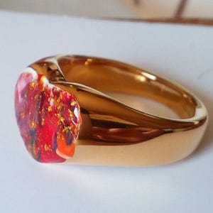 May include: A gold-colored ring with a vibrant, colorful stone. The stone features a mix of red, orange, and gold hues, creating a unique and eye-catching design. The ring's band has a smooth, polished finish.