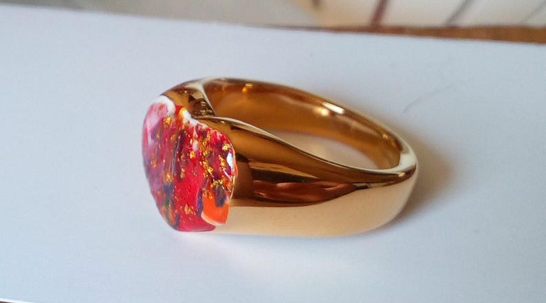 May include: A gold-colored ring with a vibrant, colorful stone. The stone features a mix of red, orange, and gold hues, creating a unique and eye-catching design. The ring's band has a smooth, polished finish.