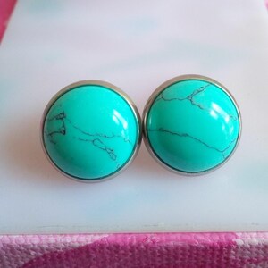 以下が含まれることがあります： 丸いスタッドイヤリングのペア。各イヤリングは、暗い静脈を持つ鮮やかなターコイズストーンを特徴とし、銀色の金属ベゼルにセットされています。イヤリングは白い表面に表示されています。