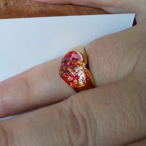 May include: A gold-colored ring with a heart-shaped, colorful resin stone. The stone features red, orange, and gold glitter accents. The ring is worn on a finger.