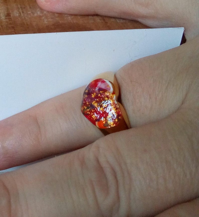May include: A gold-colored ring with a heart-shaped, colorful resin stone. The stone features red, orange, and gold glitter accents. The ring is worn on a finger.