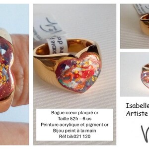 May include: A gold-plated ring with a heart-shaped face. The heart features a colorful abstract painting with red, orange, and gold accents. The ring is a unique piece of wearable art.