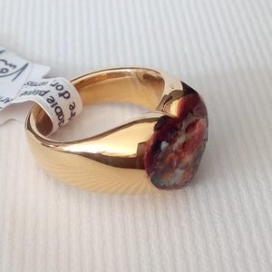 May include: A gold-colored ring with a rounded band and a colorful, abstract design on the face. The ring has a smooth, polished finish. A white tag with text is attached to the ring.
